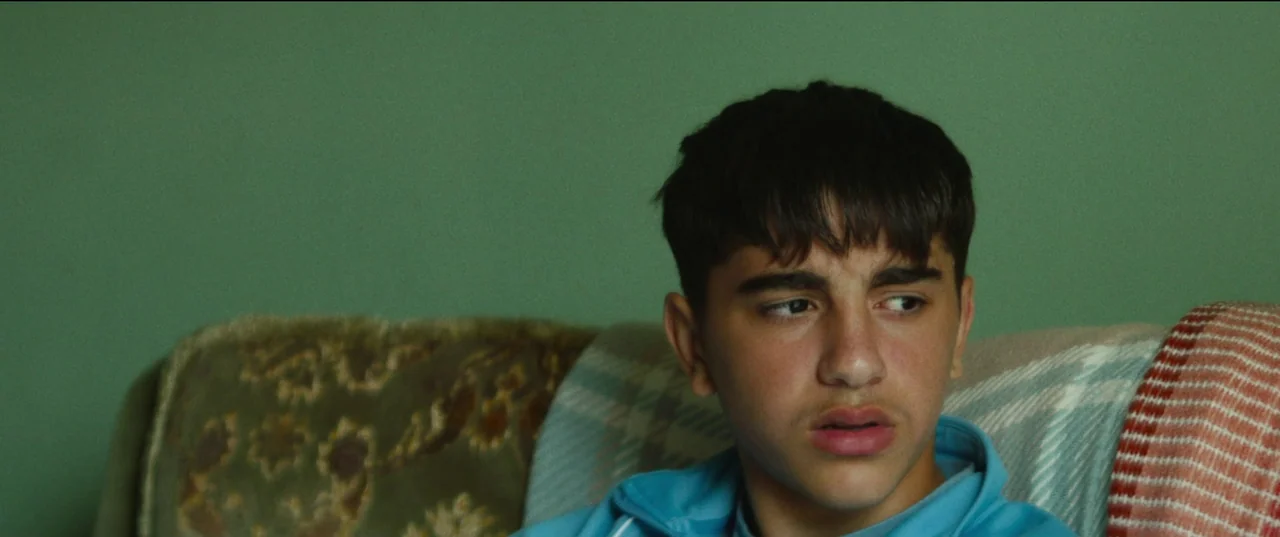Scrapper (2023) movie still: closeup — A person sits against a solid sage green wall in a furnished room. The subject, a young man, looks off-frame…