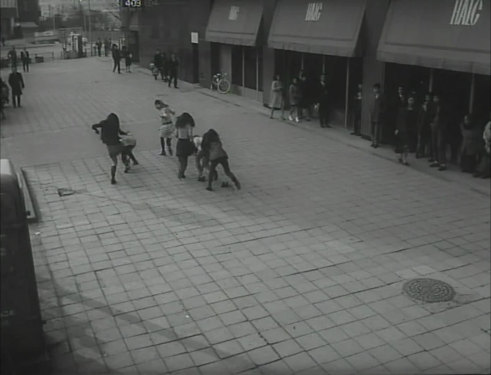 Funeral Parade of Roses (1969) movie still: wide — A paved pedestrian plaza features long buildings with striped awnings in the background. Several…