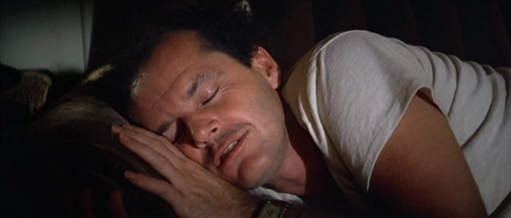 The Fortune (1975) movie still: closeup — A man sleeps in a dimly lit room against a dark surface. He rests his head upon his hand while positioned…