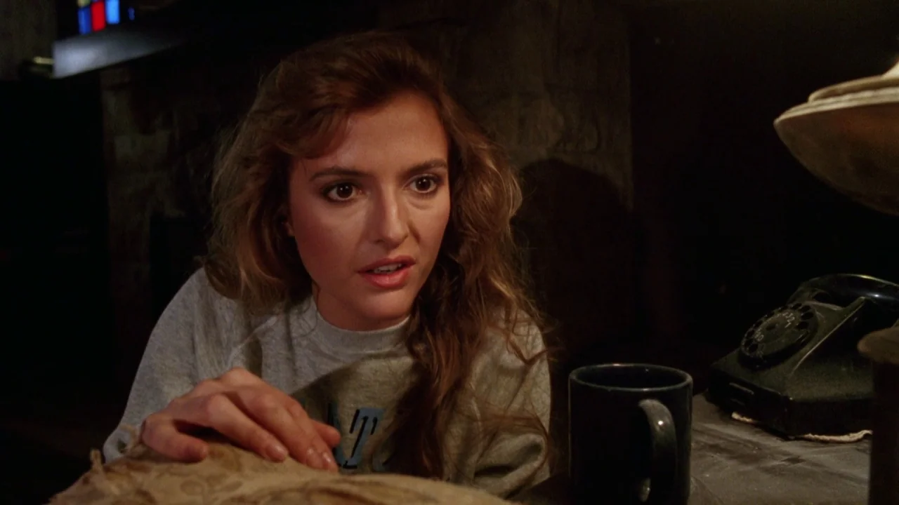 Bride of Reanimator (1989) movie still: medium — A dimly lit room features a woman leaning over a desk surface. A rotary telephone and a dark mug…