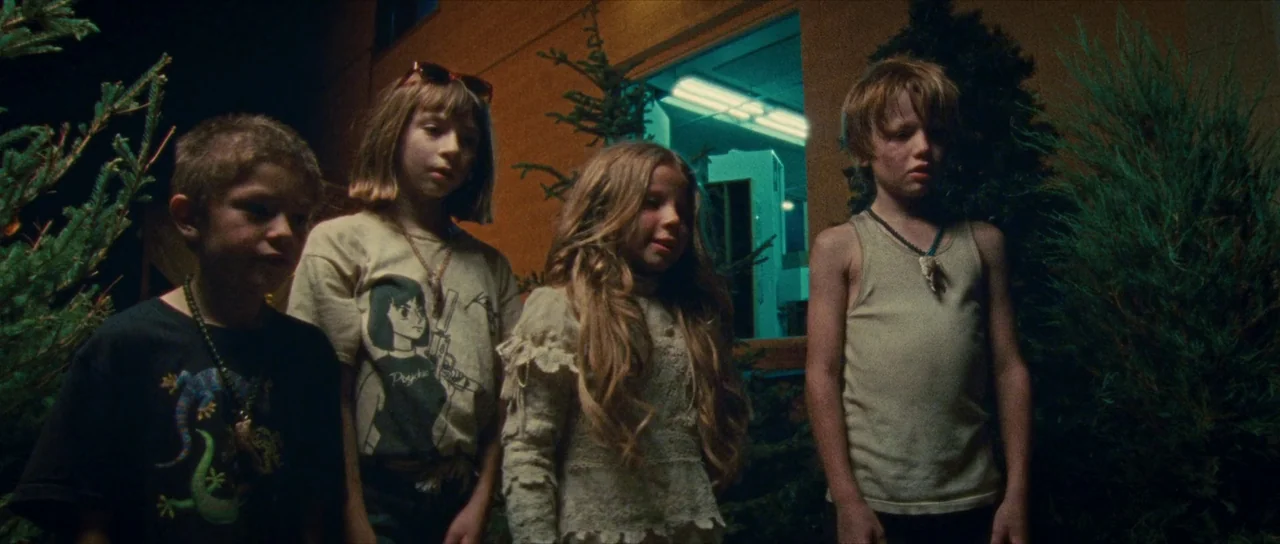 Riddle of Fire (2023) movie still: fullbody — Four children stand together outdoors at night near dense evergreen trees. A bright cyan-lit window…