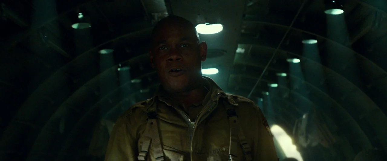 Overlord (2018) movie still: medium — The interior of a military cargo plane has rows of overhead recessed lights. A man in a tan tactical jacket…