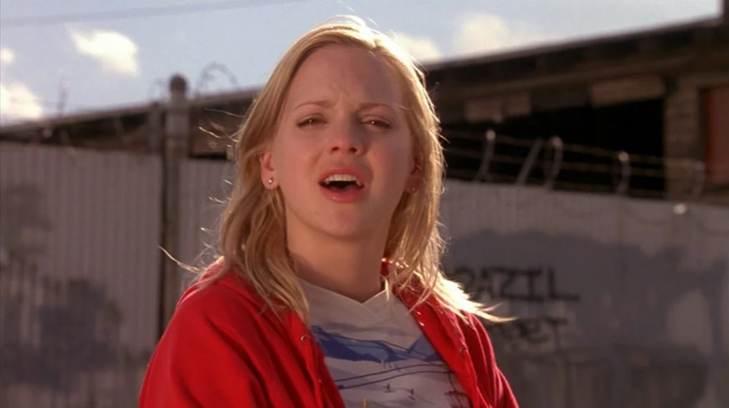 Smiley Face (2007) movie still: medium — An industrial outdoor area with a corrugated metal fence and barbed wire in background. A blonde woman in a…