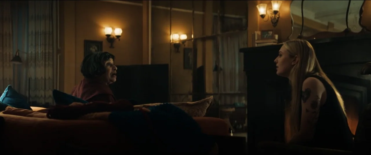 Vicious (2025) movie still: medium — A dimly lit room contains an orange couch and a brick fireplace. Two women sit facing each other engaged in a…
