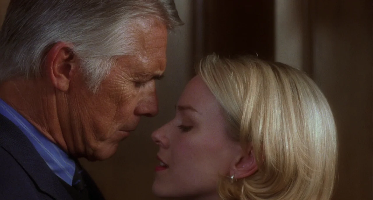 Mulholland Drive (2001) movie still: medium — Two individuals stand in an interior space with warm lighting and wooden wall textures. An older man…