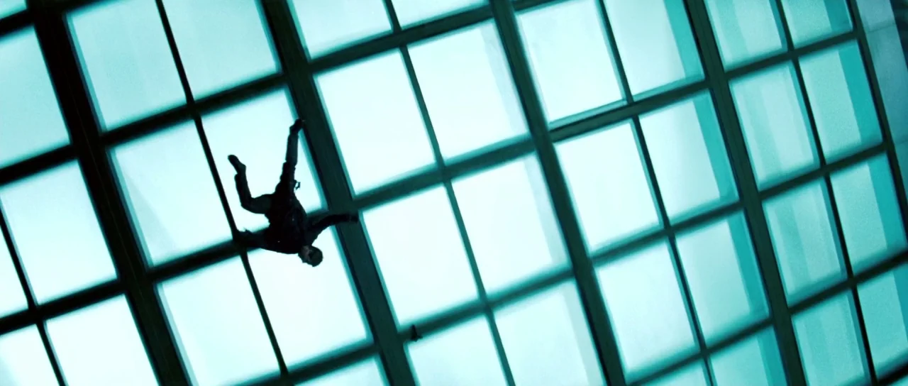Mission: Impossible III (2006) movie still: wide — A man crawls across a large, grid-patterned glass surface lit from behind. The bright blue sky is…