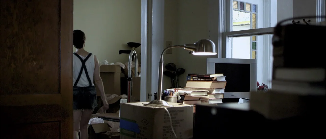 Night People (1954) movie still: medium — A dimly lit room contains stacked moving boxes and a desk with scattered books. A woman in a white tank…