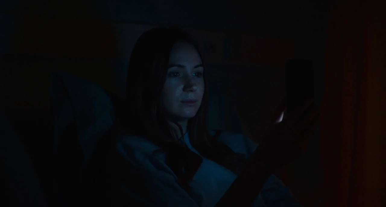 Dual (2022) movie still: closeup — A dimly lit, dark space shows a woman illuminated by her own smartphone. She sits in a position suggesting…
