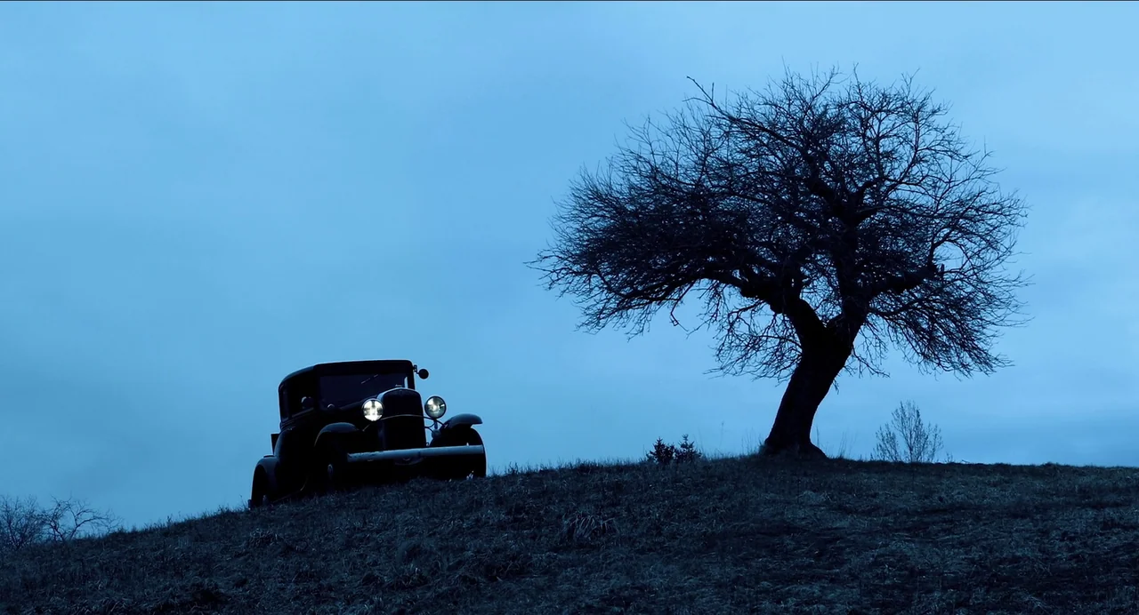 Where The Devil Roams (2023) movie still: wide — A dark vintage sedan sits atop a grassy hill during the blue hour. A large leafless tree stands…