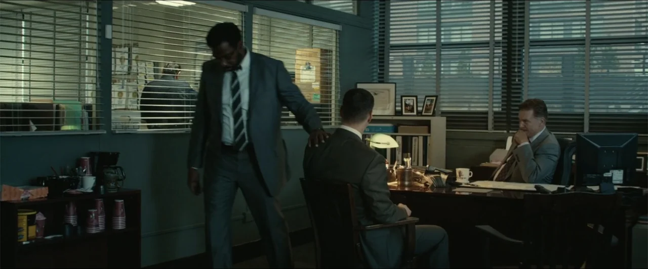 Exposed (2016) movie still: medium — A cluttered administrative workspace features metal venetian blinds covering windows on both walls. Two men in…