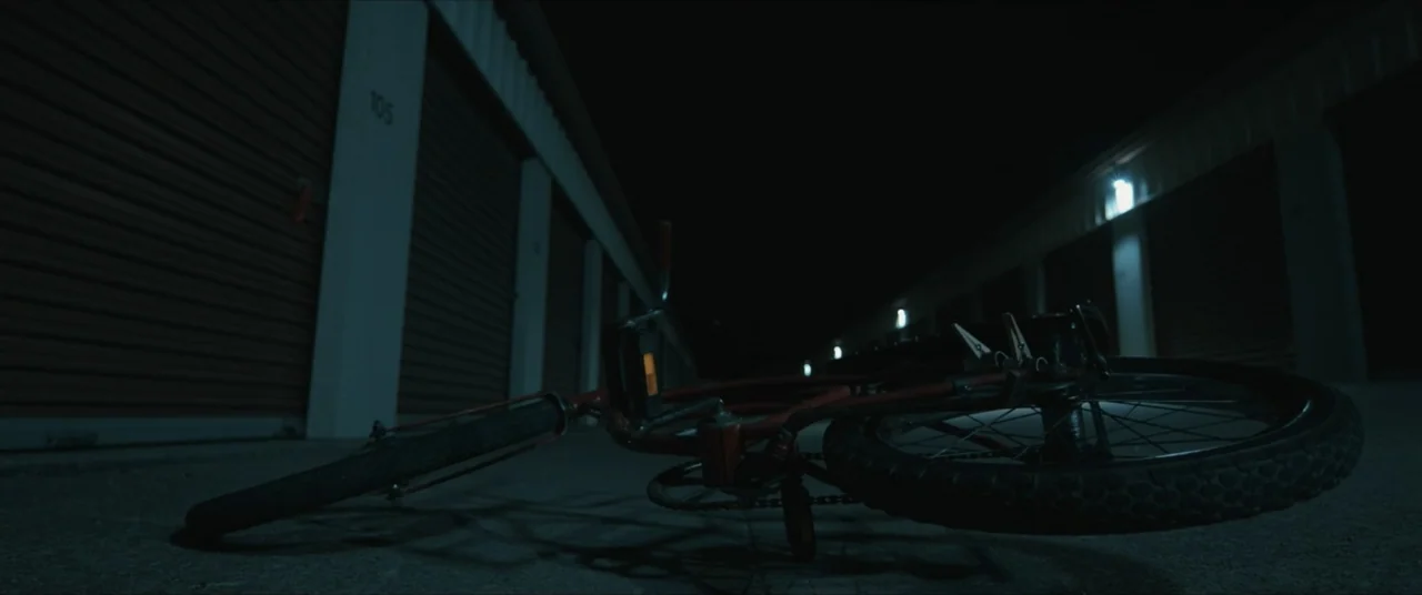 Possessions (2024) movie still: medium — An outdoor storage unit facility at night features rows of locked metal rolling doors. A red bicycle lies…
