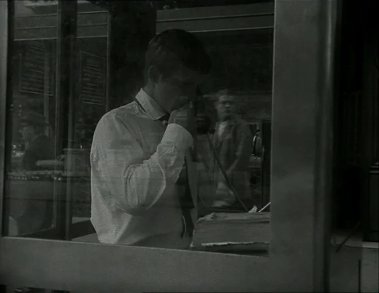 À Bout De Souffle (Breathless) (1960) movie still: medium — A man stands inside a glass telephone booth located on a busy city street. He holds a…