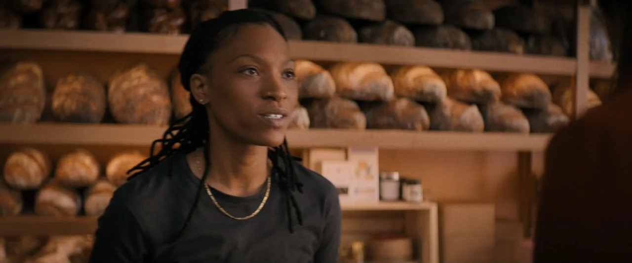 Something from Tiffany's (2022) movie still: medium — A small shop features wooden shelves stacked with numerous artisan bread loaves. A woman…
