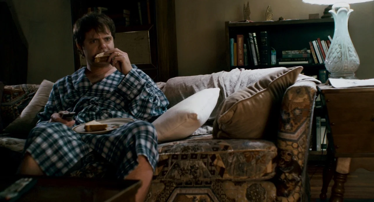 Super (2010) movie still: medium — A cluttered interior space with patterned fabric upholstery and a bookshelf backdrop. A man in a bathrobe sits on…