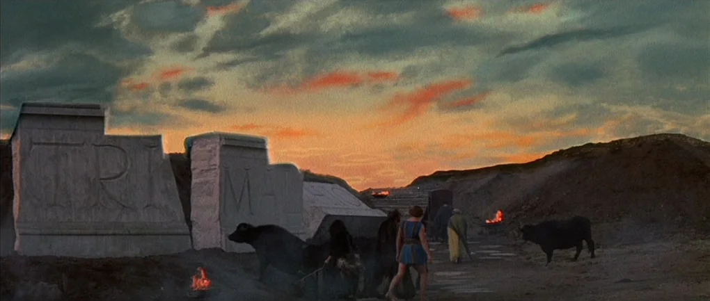 Fellini – Satyricon (1969) movie still: wide — Ancient stone blocks inscribed with letters rest on a barren dirt path at sunset. A small group of…