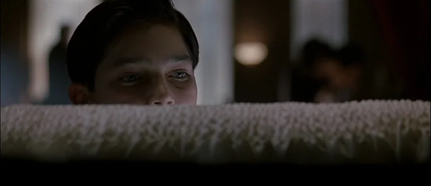 Road To Perdition (2002) movie still: closeup — A dimly lit room contains a white-lined open casket in the foreground. A teenage boy looks over the…