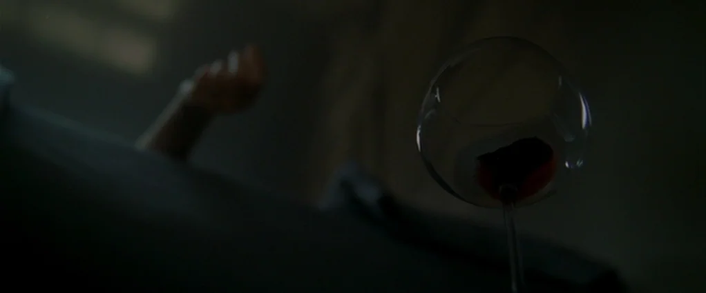 Panic Room (2002) movie still: closeup — A dimly lit, dark space contains a single glass of red wine. A blurred hand and forearm are visible resting…