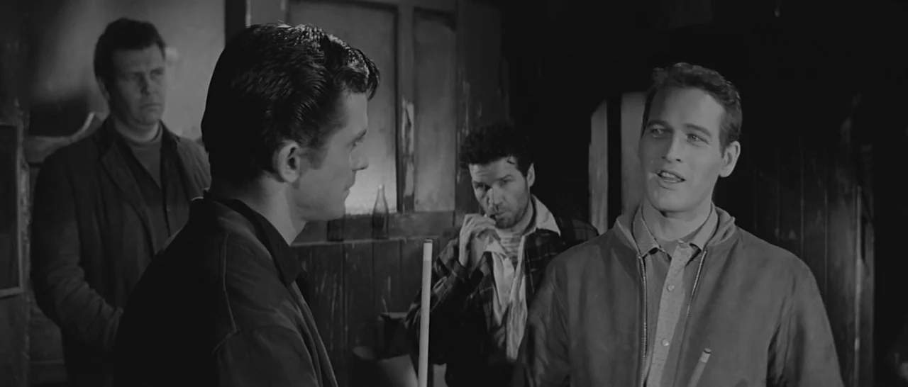 The Hustler (1961) movie still: medium — A dimly lit billiard hall with dark wood-paneled walls and a worn atmosphere. Four men stand close together…