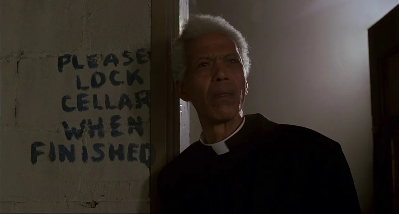 Dawn of the Dead (1978) movie still: medium — An elderly man in clerical attire stands in a dimly lit stairwell. Hand-painted text on the concrete…