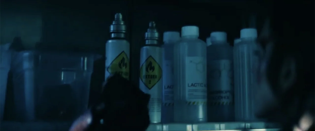 Ash (2025) movie still: medium — A dimly lit shelf in a storage area holds rows of labeled chemical containers. A person stands in the foreground,…