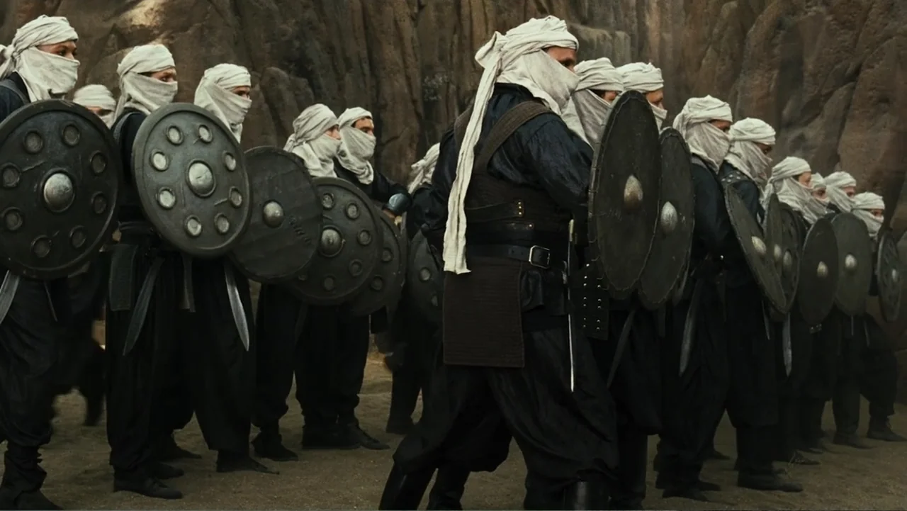Meet the Spartans (2008) movie still: medium — A group of warriors stands in a line before a sheer desert rock face. Each soldier wears a black…