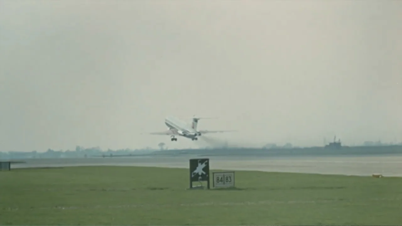 Becoming Led Zeppelin (2025) movie still: wide — A flat grassy airfield stretches toward a pale sky under an overcast day. A commercial jet airplane…