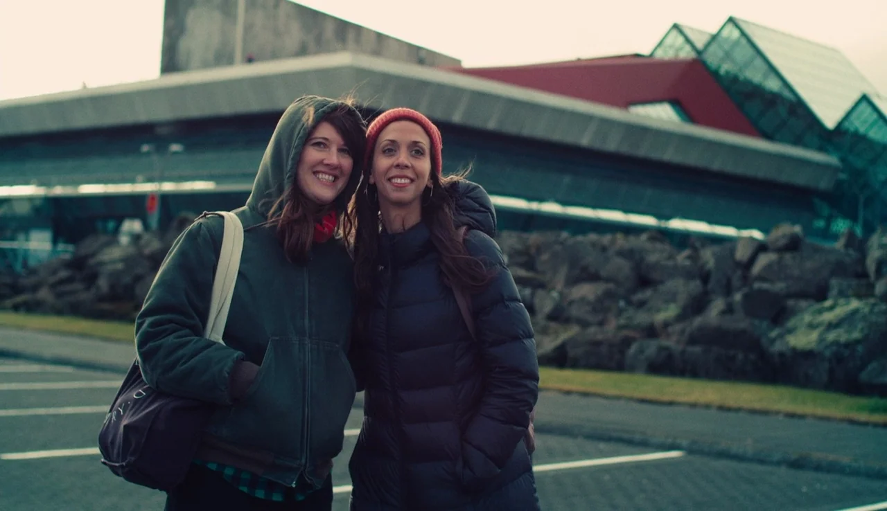 Land Ho! (2014) movie still: medium — Two women stand in a gray asphalt parking lot before a large modern building. They look toward an off-screen…