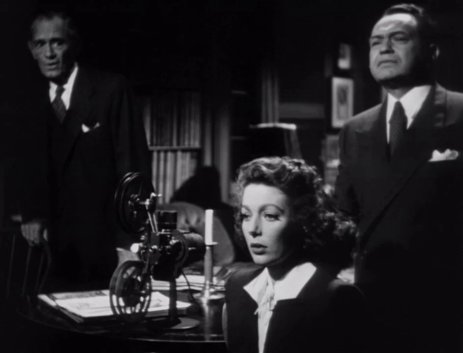 The Stranger (1946) movie still: medium — A dark, wood-paneled room features a vintage film projector on a circular table. A woman sits in the…
