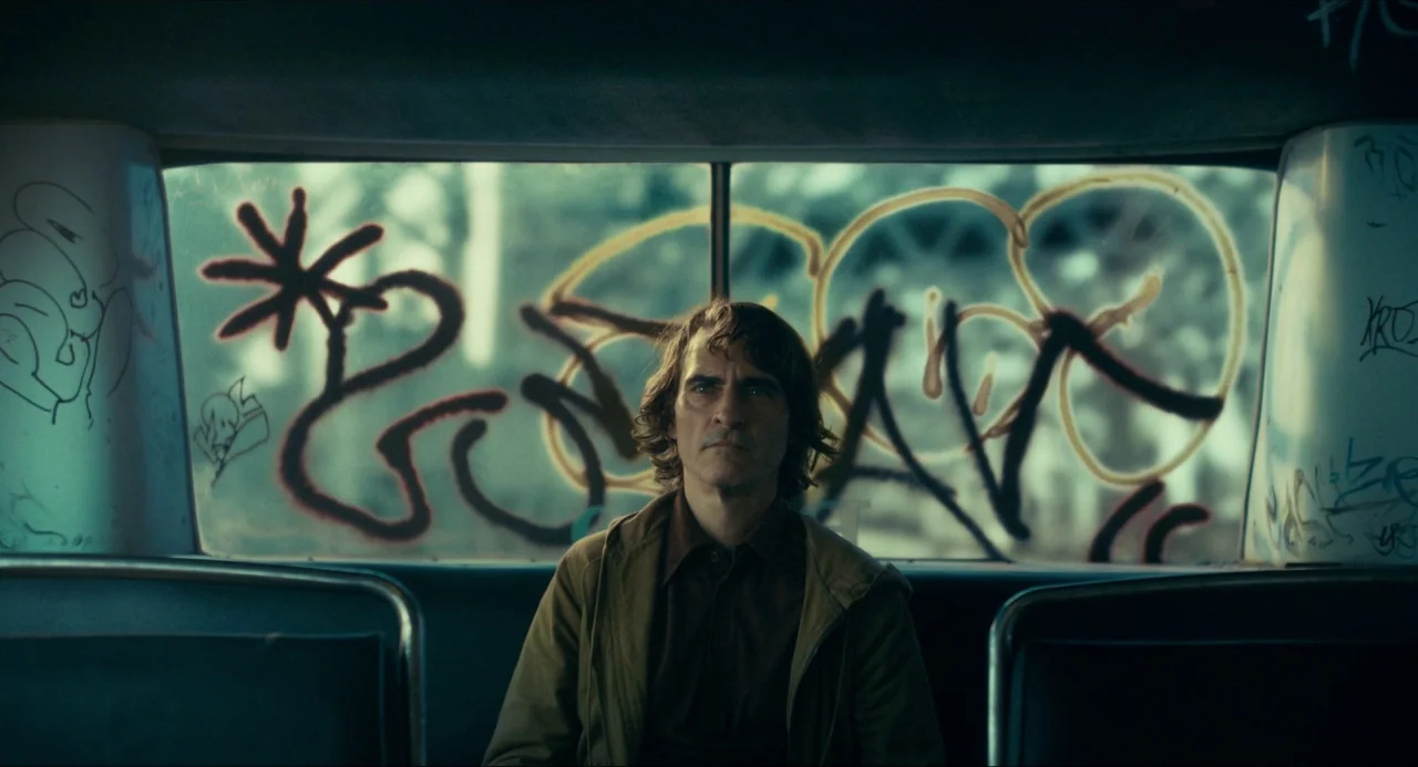 Joker (2019) movie still: medium — A man sits inside a bus with graffiti-covered windows showing blurred urban scenery outside. He stares ahead…