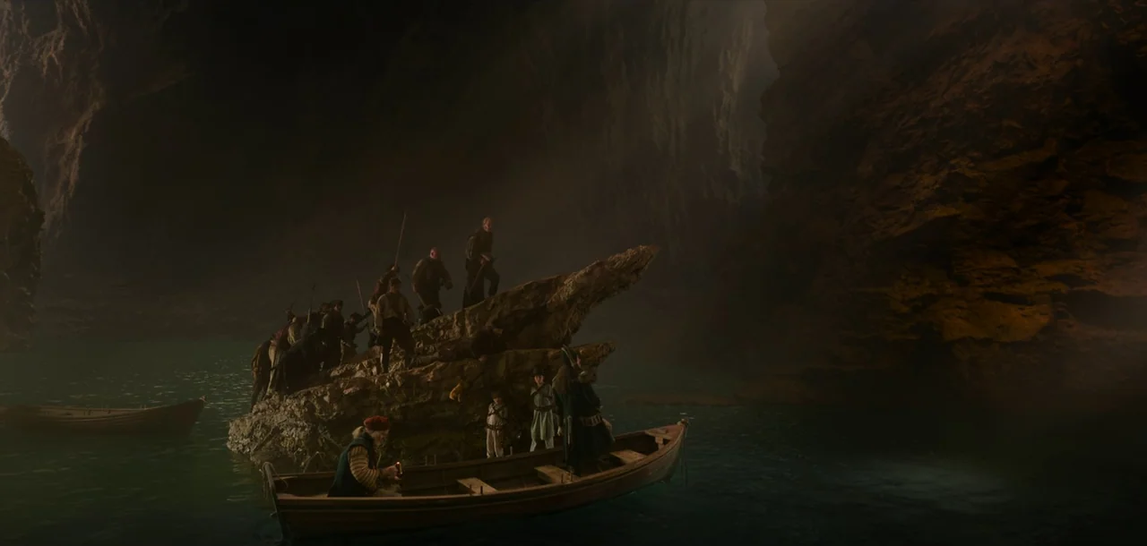 Peter Pan & Wendy (2023) movie still: wide — A dark subterranean lake surrounded by massive jagged cave walls with low light. A group of people on a…