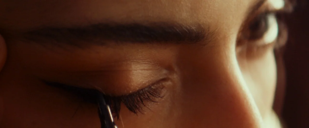 Animalia (2023) movie still: closeup — A close-up view focuses on a human eye during a makeup application session. The subject carefully applies…