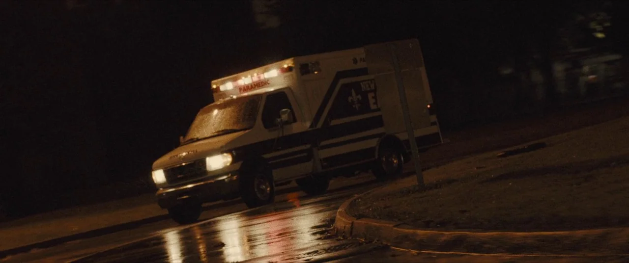 Synchronic (2019) movie still: wide — A white ambulance with dark striping sits on a dark, wet asphalt road surface. The vehicle's headlights…