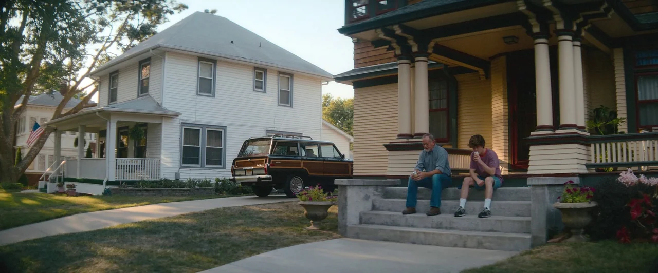 Snack Shack (2024) movie still: wide — Two suburban houses sit adjacent to each other with a wood-paneled station wagon parked nearby. An adult man…