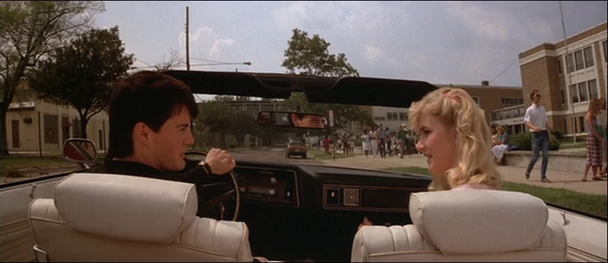 Blue Velvet (1986) movie still: medium — Two individuals sit in the front seats of a convertible parked outside a school. Other students walk across…