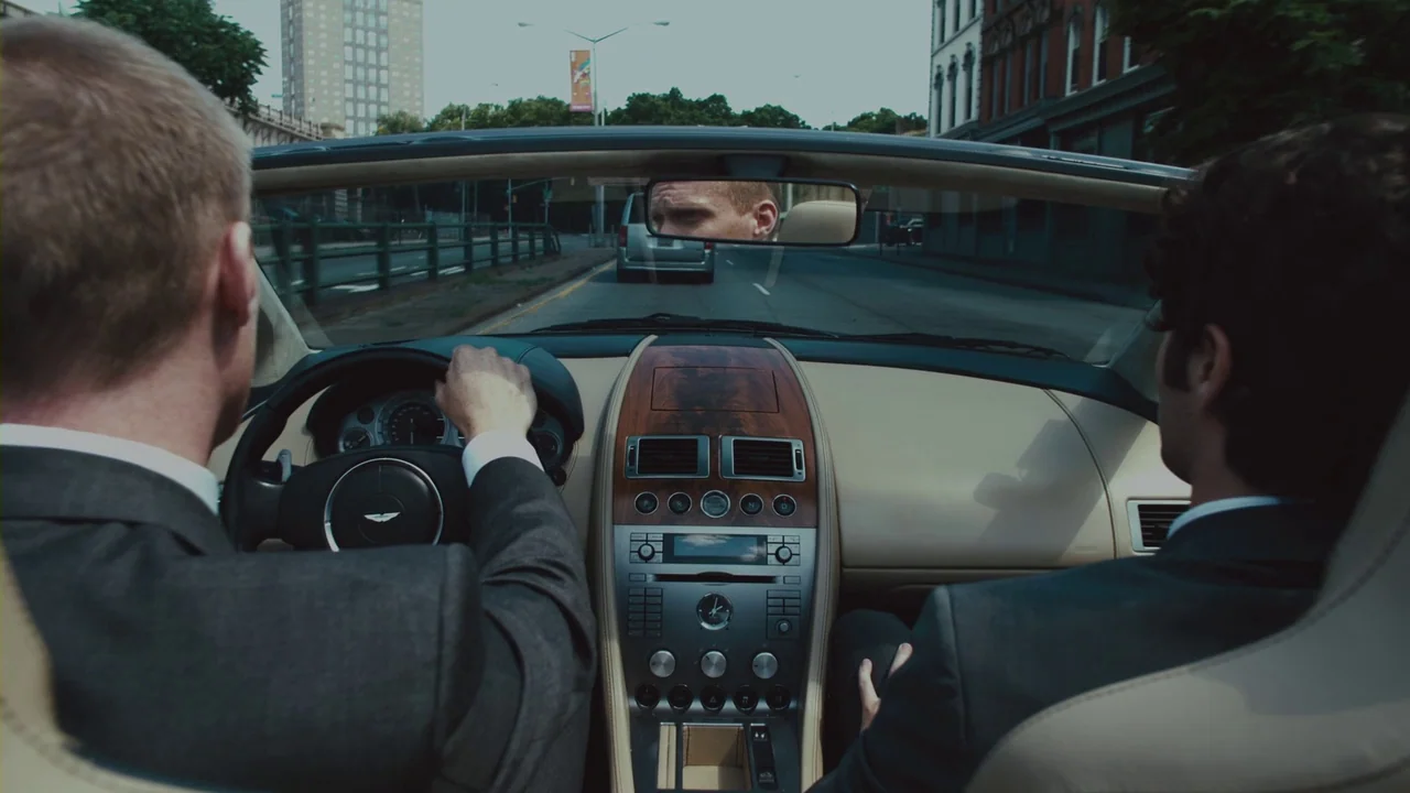 Margin Call (2011) movie still: medium — Two men in suits ride in an Aston Martin convertible on a city road. The driver's face is visible through…