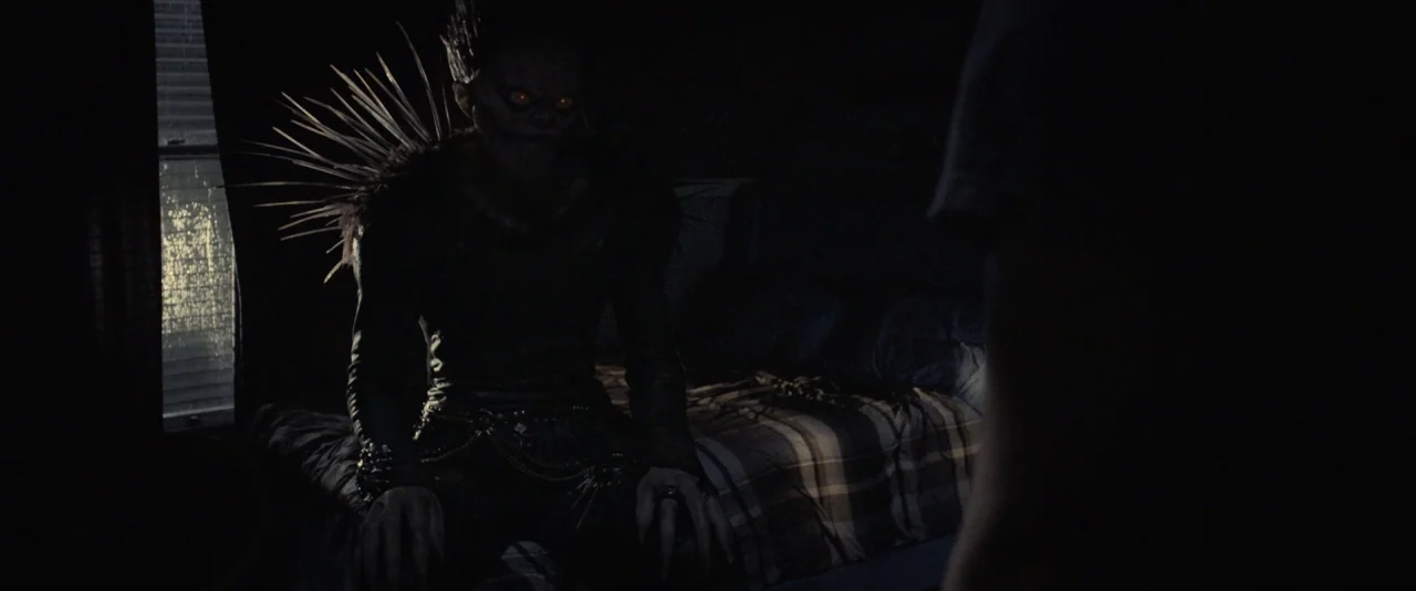 Death Note (2017) movie still: medium — A dark, dimly lit room contains a plaid-patterned mattress in the background. A supernatural entity with…