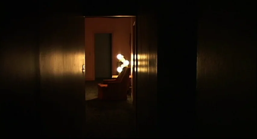 Inland Empire (2006) movie still: wide — An empty darkened room is viewed through a partially opened doorway. A lone upholstered chair stands…