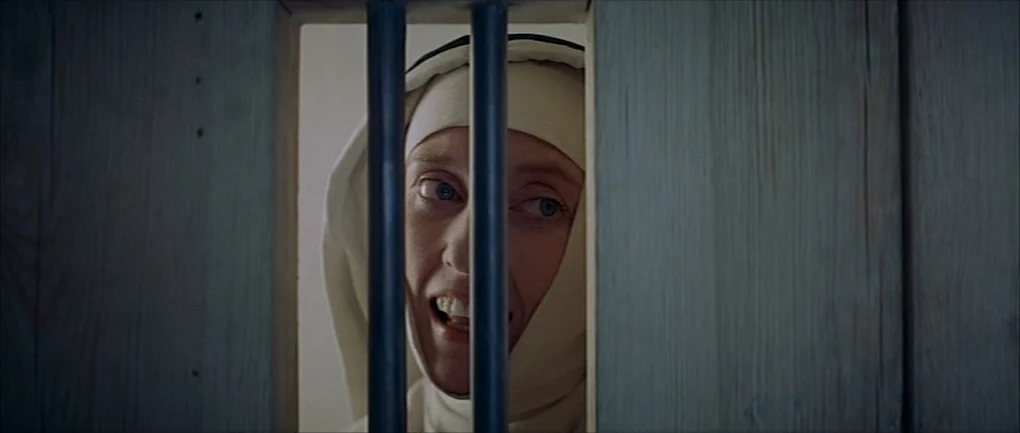 The Devils (1971) movie still: closeup — A woman dressed in a nun's habit peers through a vertical opening in wooden slats. She looks directly…