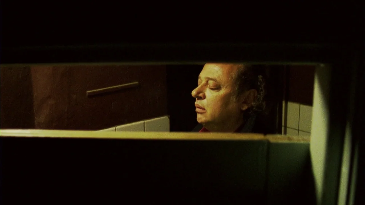 Pusher III (2004) movie still: closeup — A dimly lit room with dark walls and white wall tiles. The man stands with his eyes closed in the center of…