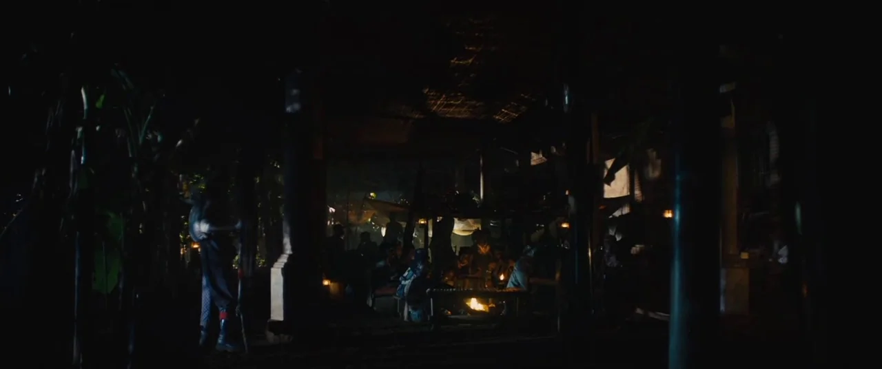 Beasts of No Nation (2015) movie still: wide — A dark, sheltered area features wooden support columns and a thatched roof structure. A central fire…