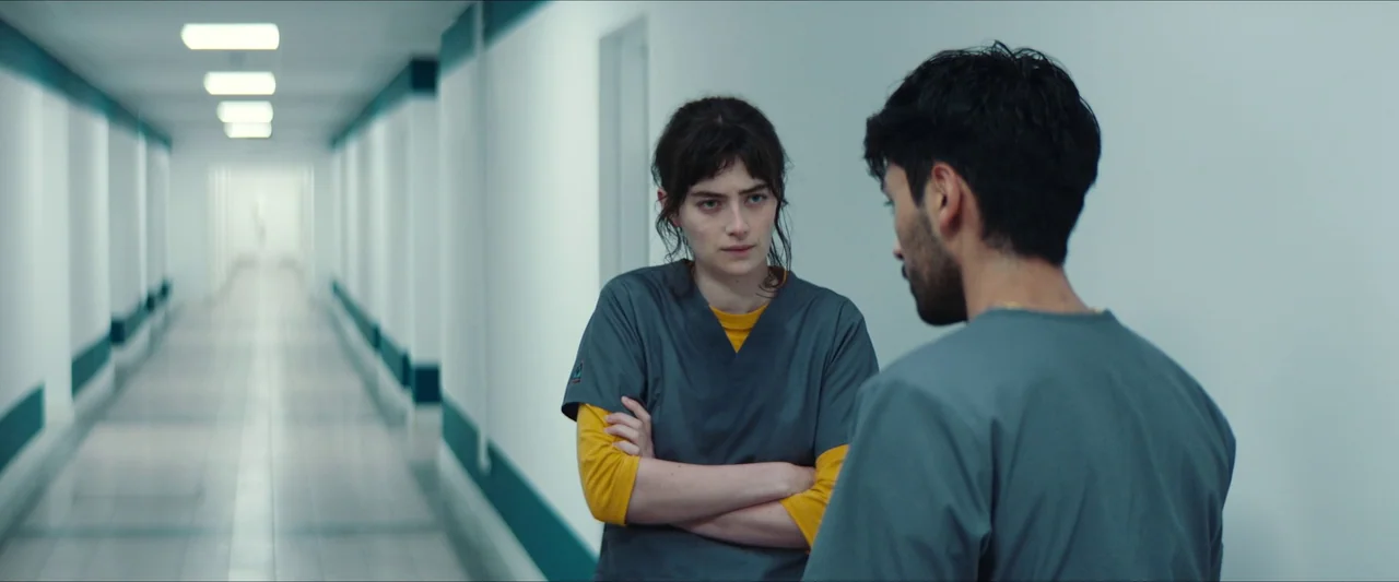 Double Blind (2023) movie still: medium — A long white institutional hallway with teal accent lines on the walls and floors. A young woman in scrubs…