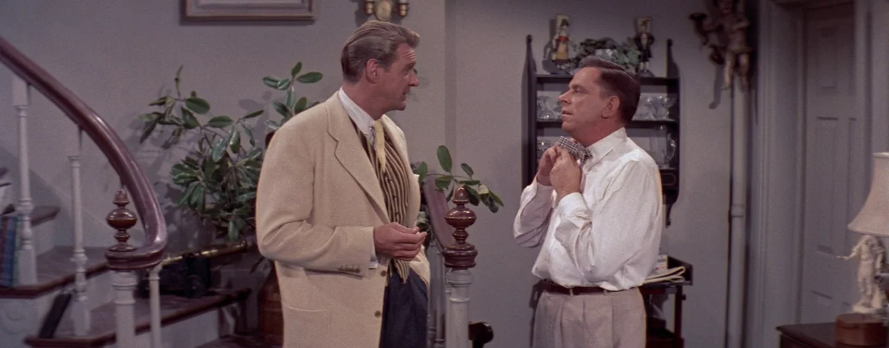 The Seven Year Itch (1955) movie still: medium — A residential hallway with light gray walls features a curved wooden staircase railing. Two men…