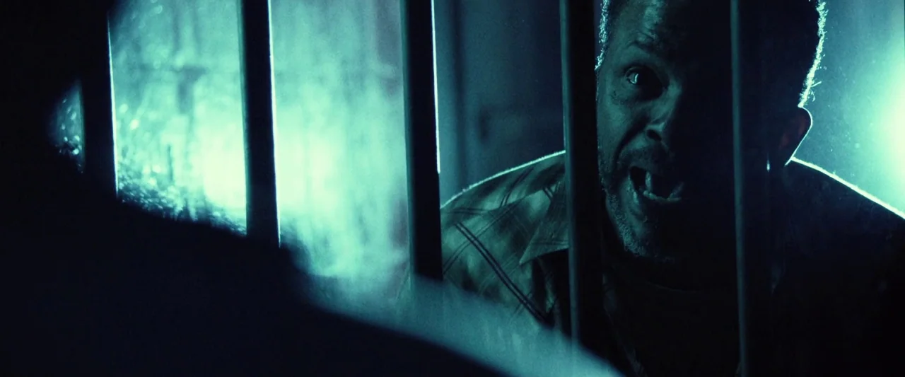 31 (2016) movie still: medium — A dimly lit space features heavy vertical metal bars obscuring a man's face. The man stands behind the metal…