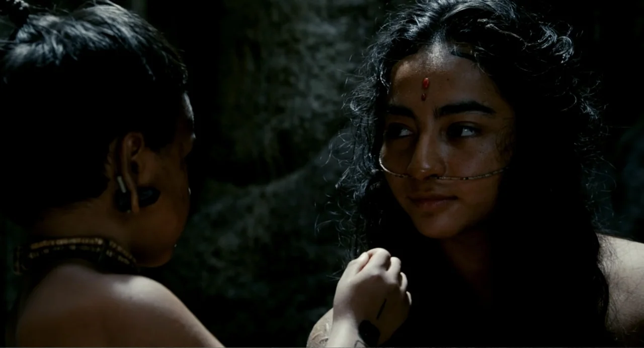 Apocalypto (2006) movie still: medium — A dark, dense jungle clearing provides a shadowed backdrop for a close encounter. An adult woman looks…