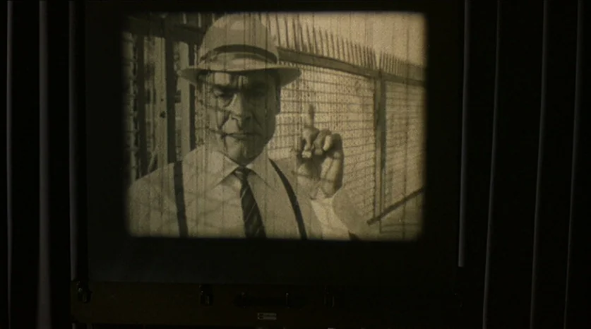 Juliet of the Spirits (1965) movie still: closeup — A man stands behind a chain-link fence on a bright, sunny day. He holds up one index finger…