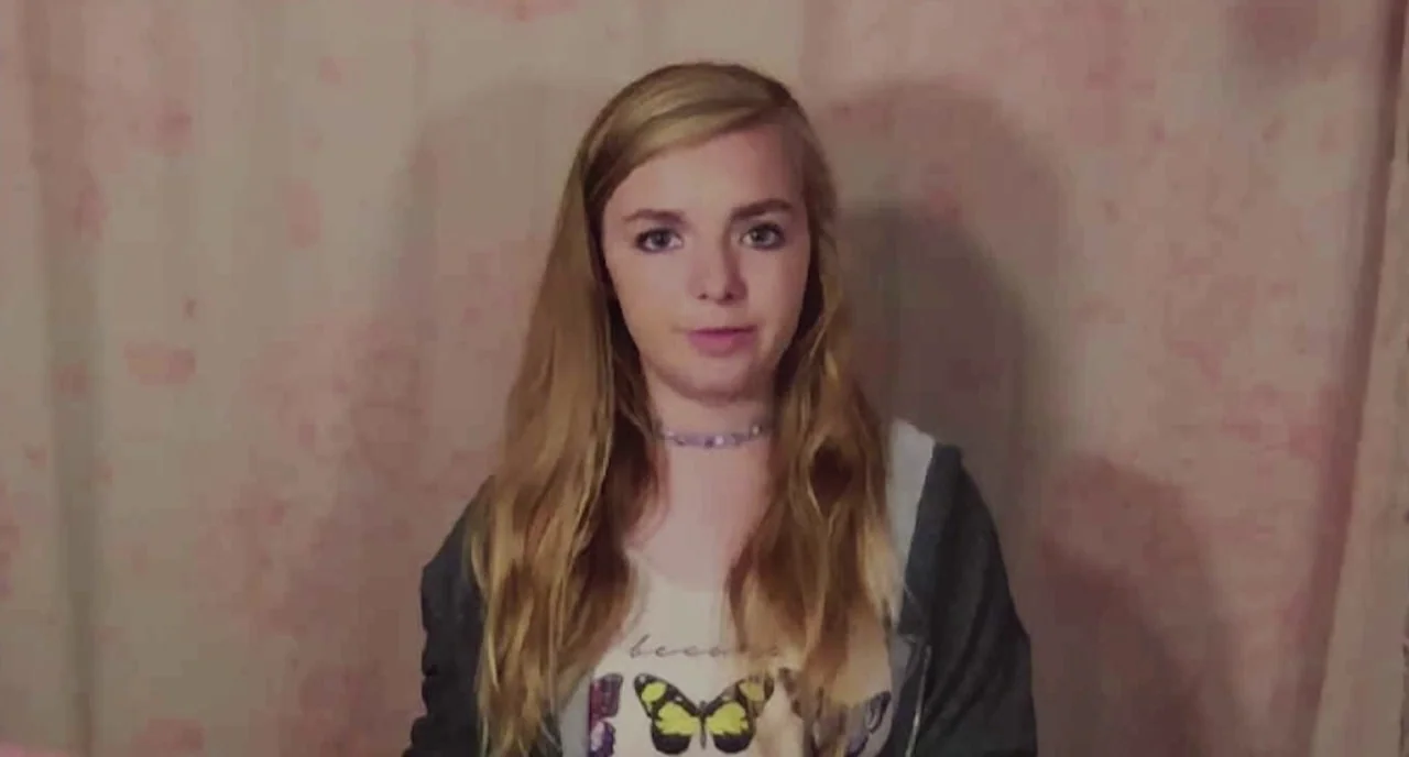 Eighth Grade (2018) movie still: medium — A teenage girl stands in front of a plain pink wall with patterned wallpaper. She speaks toward the viewer…
