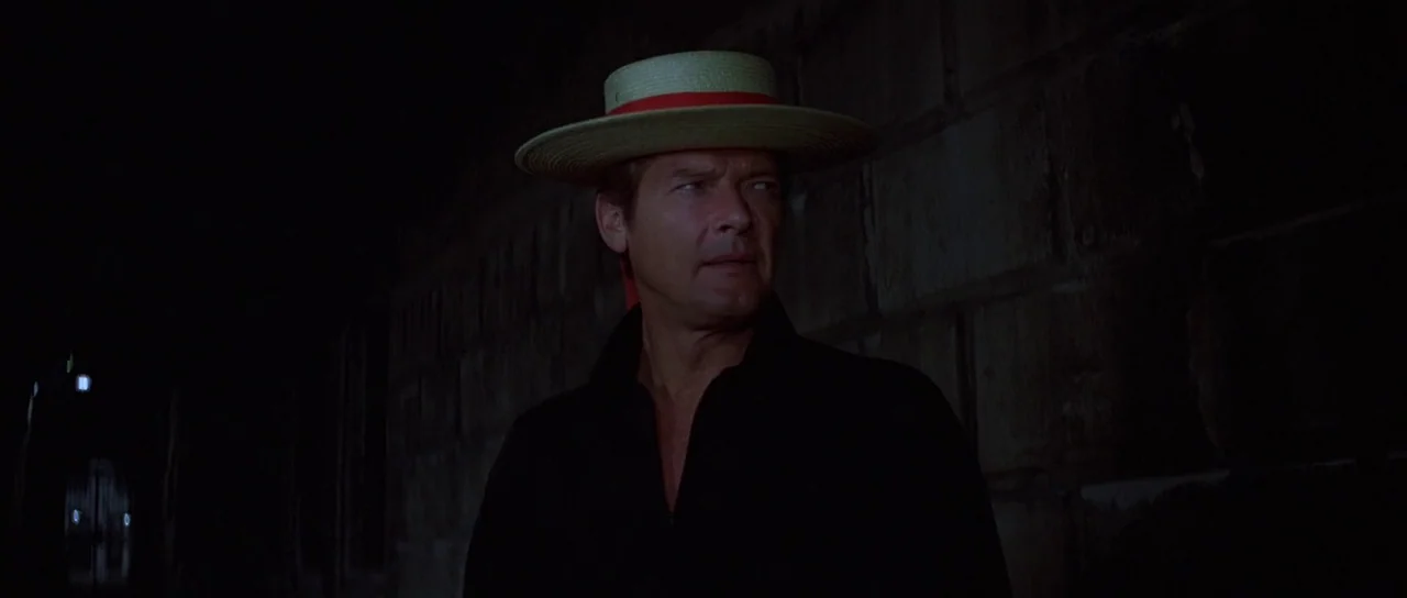 Moonraker (1979) movie still: medium — A man stands outdoors at night near a dark, block-patterned stone wall. He wears a straw boater hat and looks…