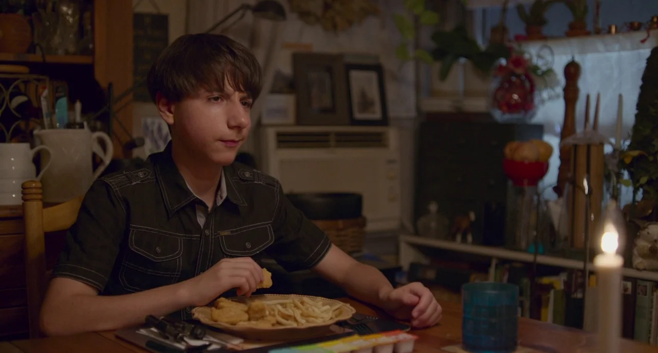 Eighth Grade (2018) movie still: medium — A cluttered dining room table holds a plate of chicken nuggets and french fries. A young boy in a black…