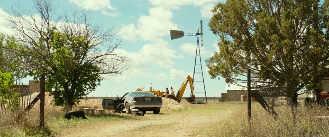 Hell or High Water (2016) movie still: wide — A rural ranch landscape features a windmill, a yellow backhoe, and a parked car. Dry vegetation…