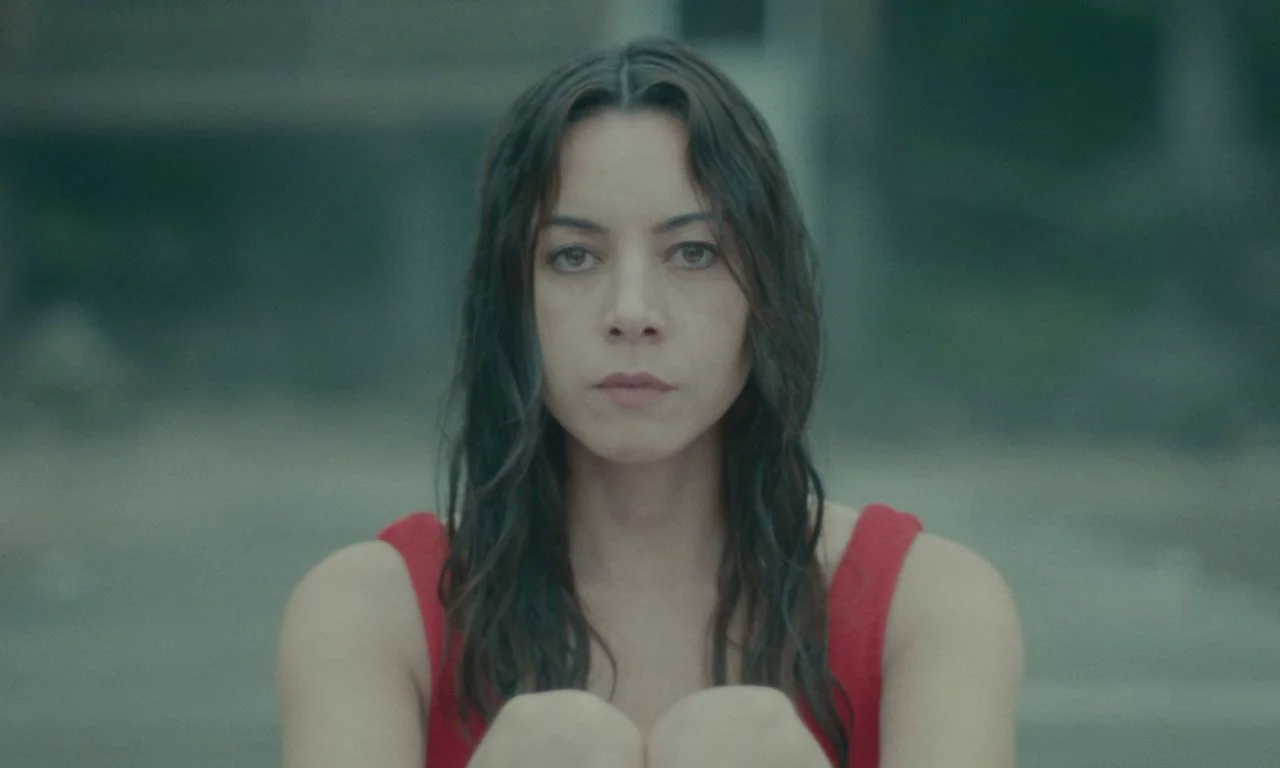 Black Bear (2020) movie still: medium — A woman sits outdoors with an out-of-focus background of muted grey tones. She looks directly forward with…
